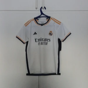  Real Madrid fotbollströja - Säljer en vit Real Madrid fotbollströja med korta ärmar och svarta detaljer. På baksidan står 'VINI JR. 20'. Perfekt för fans av laget! ¡Hala Madrid!