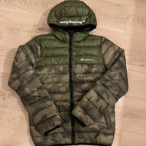 Camouflage pufferjacka från Champion - Säljer en grön och camouflagemönstrad pufferjacka från Champion.  Jackan har en huva och dragkedja framtill. Perfekt för kyliga dagar med sin quiltade design. Jackan är i nyskick. Storlek S herr. 