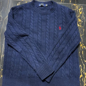 Mörkblå kabelstickad tröja från Ralph Lauren - Säljer en stilren mörkblå kabelstickad tröja från Ralph Lauren med det klassiska röda logotypbroderiet på bröstet. Tröjan har långa ärmar och en rund halsringning. Perfekt för kyliga dagar! Priset går att diskuteras vid snabb affär 