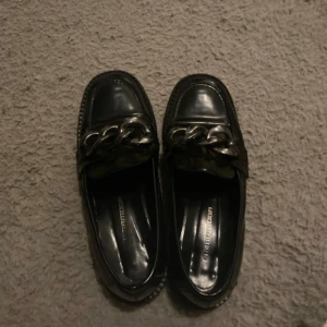 Svarta loafers med kedjedetalj - Snygga svarta loafers med en iögonfallande kedjedetalj på ovansidan. Perfekta för en stilren look. Skorna har en klassisk design med rundad tå och låg klack.