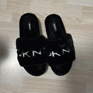Svarta tofflor från DKNY - Mysiga svarta tofflor från DKNY med mjukt pälsmaterial och bred rem över foten. Perfekta för att hålla fötterna varma och bekväma inomhus. DKNY-loggan är tydligt synlig på remmen. PRIS KAN DISKUTERAS!