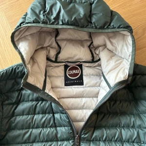 Grön colmar jacka - Säljer en snygg grön quiltad jacka från Colmar. Jackan har en praktisk huva och dragkedja framtill. Perfekt för kyligare dagar med sin pufferdesign. Två fickor med dragkedja på sidorna för extra förvaring.