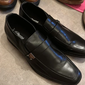 Svarta loafers från Stepside Comfort - Snygga svarta loafers från Stepside Comfort i skinn. De har en elegant design med en dekorativ rem över vristen. Perfekta för en stilren look.