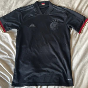 Svart fotbollströja från Adidas - Snygg svart fotbollströja från Adidas med korta ärmar och Tysklands landslagsemblem på bröstet. Tröjan har tre stjärnor ovanför emblemet och subtila ränder på axlarna. Perfekt för fotbollsfans!
