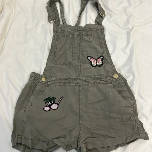 Grå hängselshorts med broderade detaljer - Säljer ett par grå hängselshorts med supercoola broderade detaljer! De har en söt fjäril och en palm med solglasögon på framsidan. Perfekta för en avslappnad sommarlook. Shortsen har justerbara axelband och praktiska fickor. De är i bra skick och redo för nya äventyr!