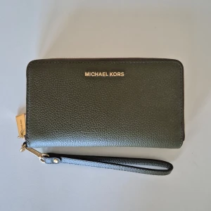 Plånbok Michael Kors - Plånbok från märket Michael Kors. Oanvänd och mycket god skick. Flera fickor för kort mm. Handtag för handen. Fin accessoar att bära bara som den är. Olivgrön färg.  18 cm lång och 10 cm hög