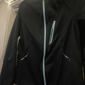 Svart vindjacka från Crivit - Säljer en svart vindjacka från Crivit med ljusblå dragkedja och detaljer. Perfekt för blåsiga dagar och utomhusaktiviteter. Jackan har en sportig look och är i bra skick.
