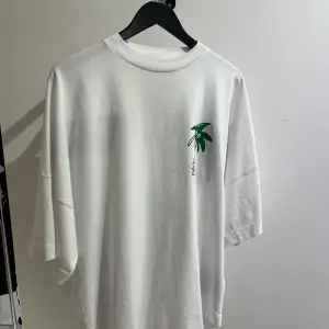 Säljer en snygg vit t-shirt från Palm Angels med ett coolt palmtryck i grönt och brunt på bröstet och ryggen. T-shirten har en avslappnad passform och korta ärmar, perfekt för sommardagar. Den är i nyskick och redo för nya äventyr!