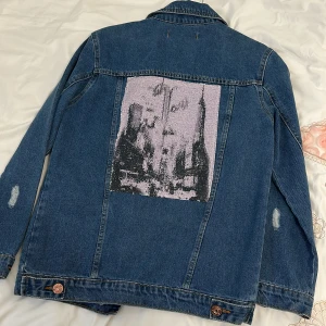 Jeansjacka från Viola Jeans - Säljer en snygg jeansjacka från Viola Jeans. Den har en cool sliten look med flera slitningar och en unik design på ryggen. Jackan har knappar framtill och två bröstfickor med lock. Perfekt för en avslappnad stil!