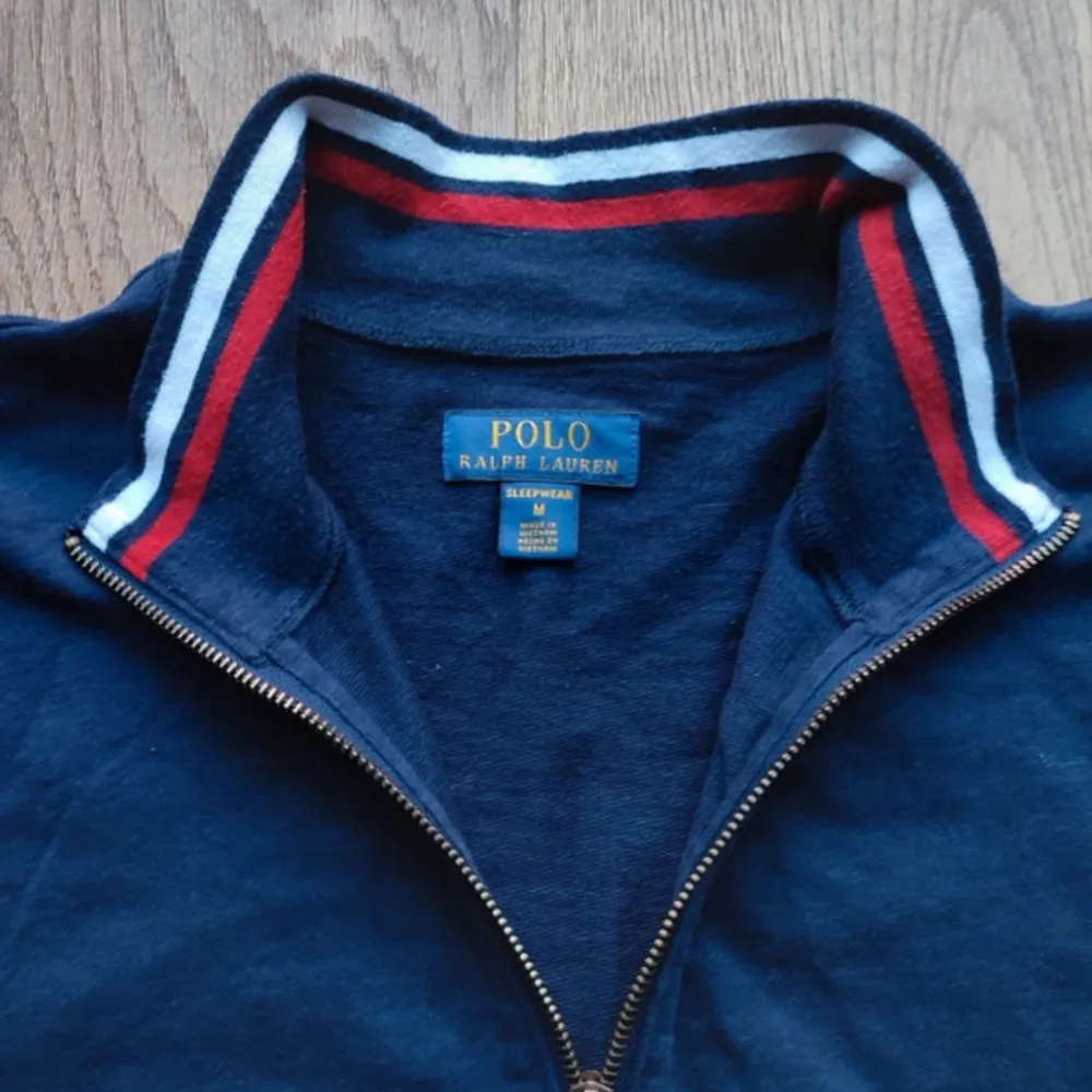 Säljer en snygg mörkblå tröja från Polo Ralph Lauren. Den har en dragkedja framtill och en stilren design med röd logga på sidan. Kragen har en cool randig detalj i rött och vitt. Perfekt för en avslappnad stil!. Tröjor & Koftor.