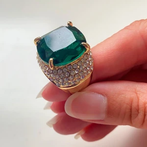 Guldring med stor grön sten och strass - Säljer en riktigt iögonfallande guldfärgad ring med en stor grön sten i mitten. Ringen är dekorerad med massor av små glittrande strass runt om, vilket ger den en lyxig och glamorös känsla. Perfekt för att dra uppmärksamhet på festen! Okänd metall 