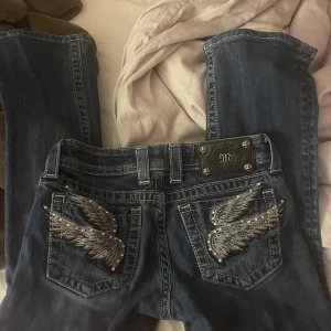 Blå jeans med vingbroderi - Snygga blå jeans med unikt vingbroderi och strassdetaljer på bakfickorna. De har en bootcut-stil och är designade i USA. Perfekta för dig som vill sticka ut med en cool detalj på dina jeans.