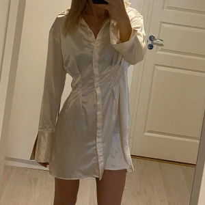 Vit skjortklänning från Divided - Snygg vit kortklänning från Divided i ett glansigt material. Klänningen har långa ärmar och en klassisk krage. Perfekt för en stilren look. ✨Stlk 38