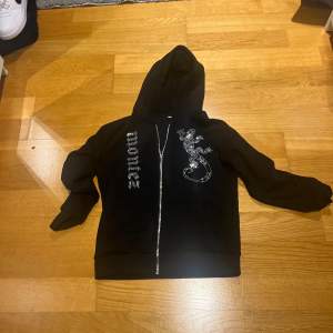 Säljer en svart hoodie med dragkedja och en glittrig ödla på framsidan. Texten 'montez' i glittriga bokstäver längs ena sidan. Perfekt för en unik och stilren look.