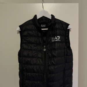 Svart dunväst från Emporio Armani - Snygg svart dunväst från Emporio Armani med EA7-logga på bröstet. Västen har en dragkedja framtill och är quiltad för extra stil och värme. Perfekt för kyligare dagar när du vill hålla stilen.