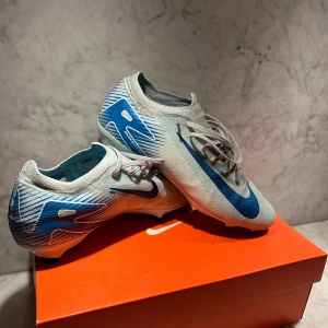 Snygga fräscha fotbollsskor från Nike  - Snygga fotbollsskor från Nike i en stilren grå färg med blå detaljer. Modellen heter ZM VAPOR 16 PRO FG. Andänder inte för det är för små. Box ingår. Nypris: 1995 kr