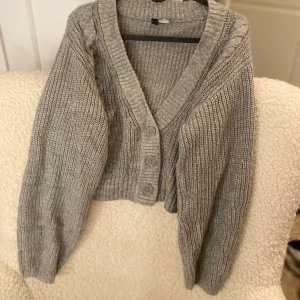 Grå stickad kofta - Mysig grå stickad kofta med knappar framtill och ett klassiskt flätmönster längs ärmarna. Perfekt för kyliga dagar och ger en stilren look. Koftan har en v-ringad design och ribbade kanter för extra komfort.