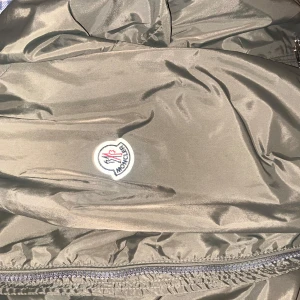 Olivgrön jacka från Moncler - Snygg olivgrön jacka från Moncler med dragkedja och huva. Jackan har Moncler-logga på bröstet och ribbade detaljer i huvan. Perfekt för en stilren look. Det är en perfekt jacka för sommaren som kommer och för våren. Det är vind jacka i storleken 2 som motsvarar storleken M den går o skanna på både koden och på märket. Perfekt skick har max använt den 5 gånger och den ser ny ut. Tyvärr inget kvitto då jag fick den i present från en familjemedlem. Först till kvarn! pris kan diskuteras 