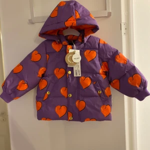 Lila pufferjacka med hjärtan från Mini Rodini - Säljer en superfin lila pufferjacka från Mini Rodini med stora orange hjärtan. Jackan har en mysig huva och stängs b knappar. Insidan är klädd i en härlig orange färg. Perfekt för att lysa upp kalla dagar! 💜🧡