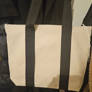 Beige shoppingväska från Rains - Snygg beige shoppingväska från Rains med svarta handtag. Perfekt för att bära med sig allt man behöver. Väskan har en stilren design och är tillverkad i ett tåligt material.