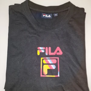 Svart t-shirt från FILA - Säljer en svart t-shirt från FILA med färgglatt logotryck på bröstet. Perfekt för en avslappnad stil. Nästan som ny. Garderobsrensning så kika gärna på övriga auktioner.