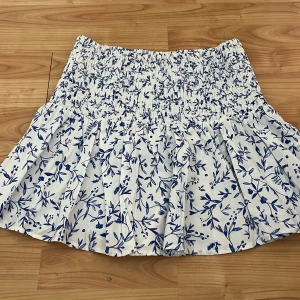 Vit och blå blommig kjol - Charmig kortkjol i vitt med blått blommönster. Kjolen har en elastisk midja som ger en bekväm passform och volangdetaljer som ger extra stil. Perfekt för varma dagar.