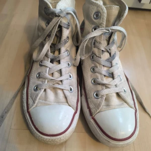 Vita Converse Chuck Taylor All Star - Klassiska vita Converse Chuck Taylor All Star sneakers med hög snörning och den ikoniska logotypen på sidan. Skorna har en vit tåhätta och röd rand längs sulan. Perfekta för en avslappnad stil.