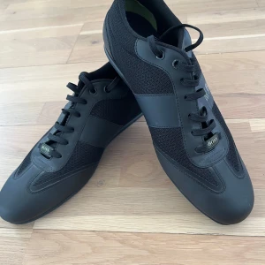 Svarta sneakers från Hugo Boss - Snygga svarta sneakers från Hugo Boss med stilren design. Skorna har snörning och en kombination av skinn och syntetmaterial. Perfekta för en modern och avslappnad stil.