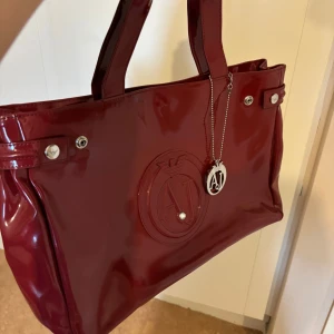 Röd axelväska från Armani Jeans - Snygg röd axelväska från Armani Jeans i glansigt material. Väskan har ett stort fack med dragkedja och ett stilrent AJ-emblem på framsidan. Perfekt för att bära med sig det viktigaste med stil.