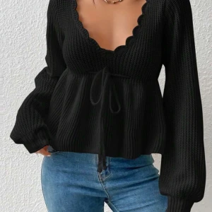 Svart stickad tröja med knyt - Snygg svart stickad tröja med djup v-ringning och knytband i midjan. Tröjan har långa ärmar och en ribbad struktur som ger en stilren look. Perfekt för kyligare dagar.