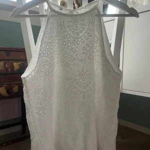 Vit ärmlös blus med broderi - Säljer en vacker vit ärmlös blus med detaljerat broderi och knytning i nacken. Perfekt för varma sommardagar eller som en elegant touch till din outfit. Blusen har ett fint mönster som ger en romantisk känsla. Den har varit en klänning innan så den är avklippt, den är dock inte så jämt avklippt!