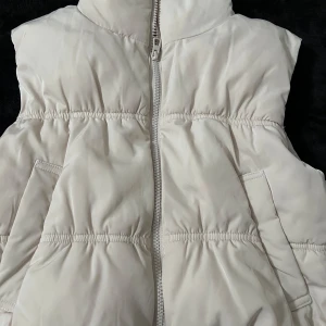 Vit dunväst från Divided - Säljer en snygg vit dunväst från Divided. Västen har en dragkedja framtill och är perfekt för kyligare dagar. Den quiltade designen ger en trendig look. Passar bra till lager-på-lager-stilen.