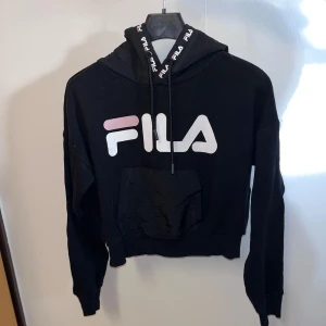 Svart hoodie från FILA - Säljer en svart hoodie från FILA med stor logga på framsidan. Hoodien har en justerbar huva med snören och en praktisk magficka. Perfekt för en avslappnad stil.