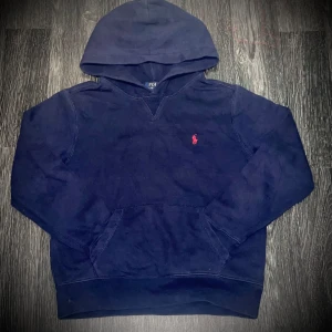 Mörkblå hoodie Ralph Lauren 10-12 år - Säljer en snygg mörkblå hoodie från Polo Ralph Lauren med det klassiska röda broderade logotypen på bröstet. Tröjan har en praktisk känguruficka och en bekväm huva. Perfekt för en avslappnad stil.