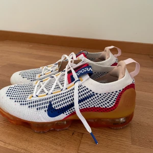 Nike Vapormax Flyknit sneakers - Snygga Nike Vapormax Flyknit sneakers i vitt med blå och röda detaljer. Skorna har en unik orange sula och är tillverkade med återvunnet material. Perfekta för en sportig look med stil.