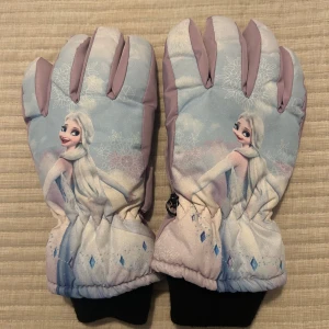 Barnhandskar med Elsa motiv - Säljer ett par barnhandskar med motiv av en populär karaktär. Handskarna är i ljuslila och blå nyanser med snöflingor och har svarta muddar. Perfekta för vintern med ett praktiskt clip för att hålla dem ihop. Passar 8-10 år. En liten skada finns.