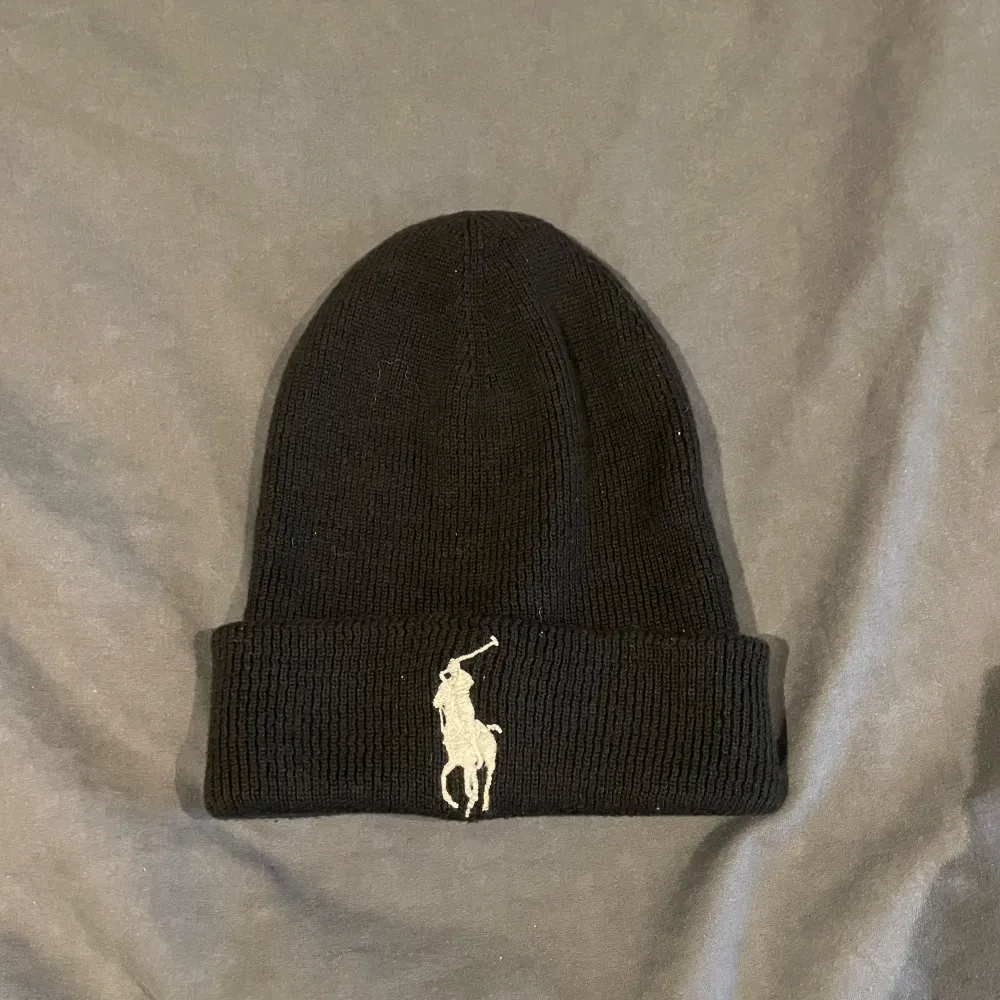 Snygg svart mössa från Polo Ralph Lauren med det ikoniska broderade logotypen framtill. Den är ribbad och perfekt för att hålla dig varm under kyliga dagar. En stilren accessoar som passar till det mesta.. Asusteet.