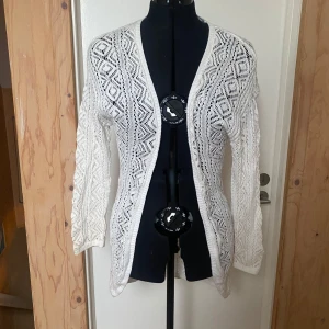 Vit stickad kofta - Säljer en elegant vit stickad kofta med ett vackert hålmönster. Koftan har långa ärmar och en öppen design framtill, perfekt för lager-på-lager-stil. Passar utmärkt till både vardag och mer uppklädda tillfällen.