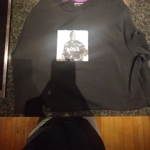 Svart tröja från Boss med tryck - Säljer en svart tröja från Boss med ett stort tryck på framsidan. Tröjan har en rund halsringning och är i en klassisk stil. Perfekt för en stilren look.