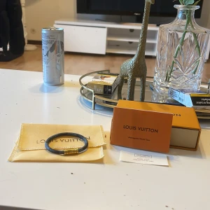 Armband från Louis Vuitton - Snyggt armband från Louis Vuitton med en elegant design. Armbandet har en mörk färg med gulddetaljer och kommer i en exklusiv förpackning. Perfekt för att ge en lyxig touch till din stil.