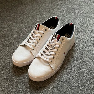 Vita sneakers från Tommy Hilfiger - Snygga vita sneakers från Tommy Hilfiger med klassisk design. Skorna har snörning och en liten logotyp på sidan. Insidan är svart med röd och vit detalj vid hälen. Perfekta för en stilren look.