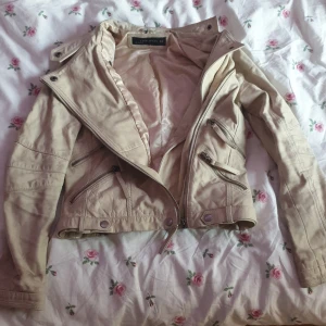 Beige skinnjacka med dragkedjor - Snygg beige skinnjacka med flera dragkedjor och knappar vid kragen. Jackan har en cool bikerstil med långa ärmar och en figurnära passform. Perfekt för att ge din outfit en edgy touch!