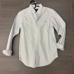 Randig skjorta från Ralph Lauren - Klassisk vit och blå randig skjorta från Ralph Lauren med broderad logga på bröstet. Ordinariepris ca 1799 kr. Helt oanvänd. Vid fler bilder eller frågor, hör av dig!