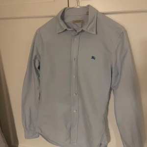Ljusblå skjorta med broderad logga - Snygg ljusblå skjorta från Burberry med en diskret broderad logga på bröstet. Skjortan har långa ärmar och knappar framtill. Perfekt för en stilren look.
