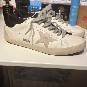 Golden goose - Snygga vita sneakers med en grå stjärna på sidan och svarta detaljer vid hälen. Skorna har grå snörning och en stilren design. Perfekta för en avslappnad stil. Har små tecken på användning men inga defekter. Tveka inte att höra av er vid frågor!