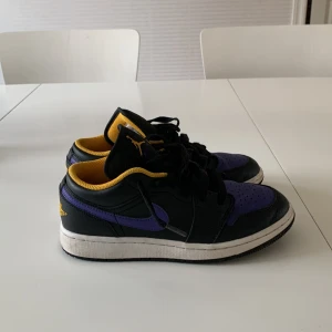 Nike Air Jordans i svart och lila - Säljer ett par Nike Air Jordans i svart och lila med gula detaljer. Skorna har snörning och en klassisk design med perforerad tå. Perfekta för dig som vill ha en stilren och sportig look.