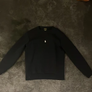Navy blue tröja från Polo Ralph Lauren - Säljer en stilren svart tröja från Polo Ralph Lauren med deras ikoniska logga broderad på bröstet. Tröjan har långa ärmar och en klassisk rund halsringning. Perfekt för en avslappnad och snygg look.