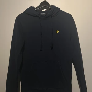 Mörkblå hoodie från Lyle & Scott - Säljer en stilren mörkblå hoodie från Lyle & Scott med en gul logotyp på bröstet. Tröjan har en klassisk design med huva och dragsko samt en praktisk magficka. Perfekt för en avslappnad look.