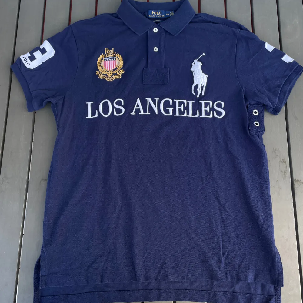 Säljer en snygg mörkblå pikétröja från Ralph Lauren. Den har en klassisk krage med knappar och är dekorerad med texten 'LOS ANGELES' i vitt på framsidan. På bröstet finns ett broderat emblem i guld och rött, och en stor vit siffra '3' på ryggen och ärmen. Perfekt för en casual men stilren look!. T-shirts.