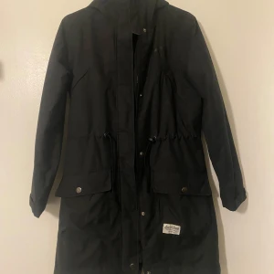 Svart parkajacka från Kari Traa - Säljer en snygg svart parkajacka från Kari Traa i storlek S. Jackan har en dragkedja och knappar framtill, samt praktiska fickor med knappar. Perfekt för höst och vår med sin långa design och justerbara midja för en bekväm passform. Märket är känt för sin kvalitet och stil, så passa på att fynda denna fina jacka!